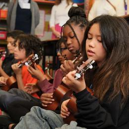 Six elementary-aged children of diverse backgrounds sit on a carpeted classroom floor, each playing a ukulele. Five adults stand behind them, observing the music lesson.