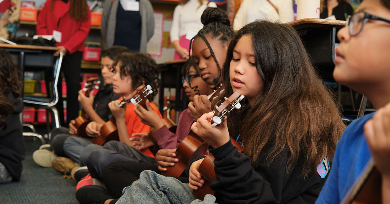 five children play ukeleles while adults look on